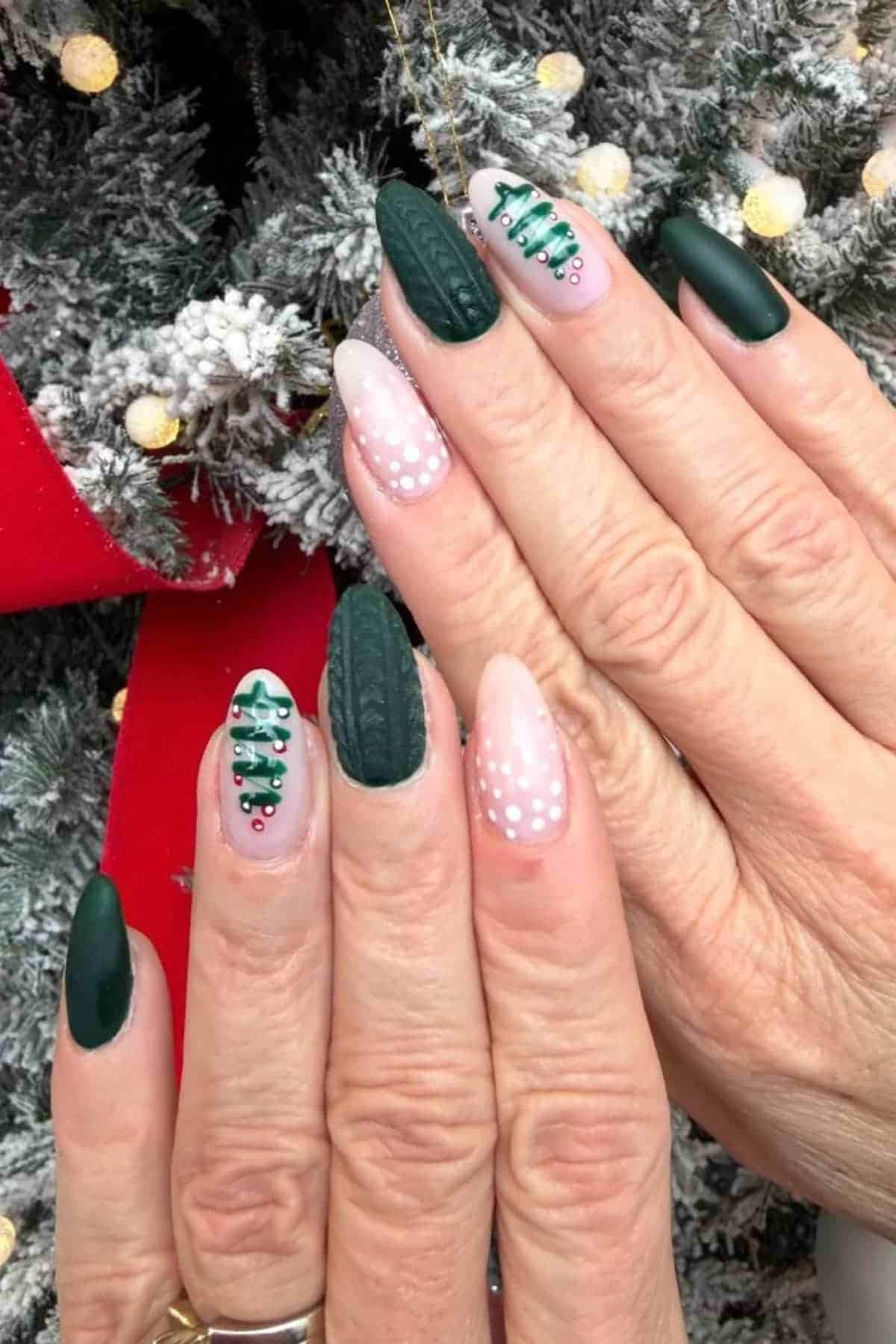 Hands with manicured nails featuring dark green, knitted texture, Christmas tree designs, and white polka dots, posed in front of a decorated, snowy artificial tree.