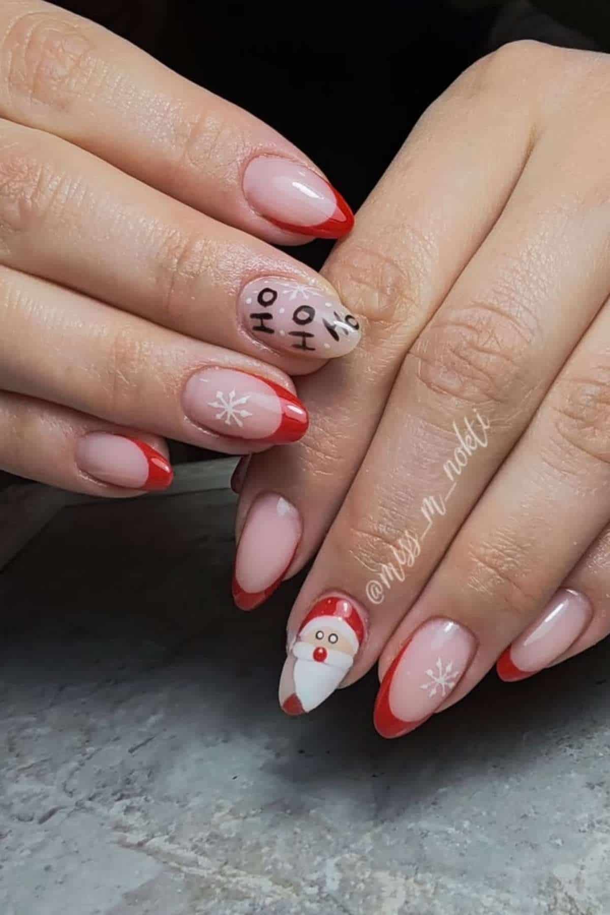 Hands with almond-shaped nails painted with red French tips, snowflake designs, and cartoon reindeer and Santa Claus faces on accent nails.