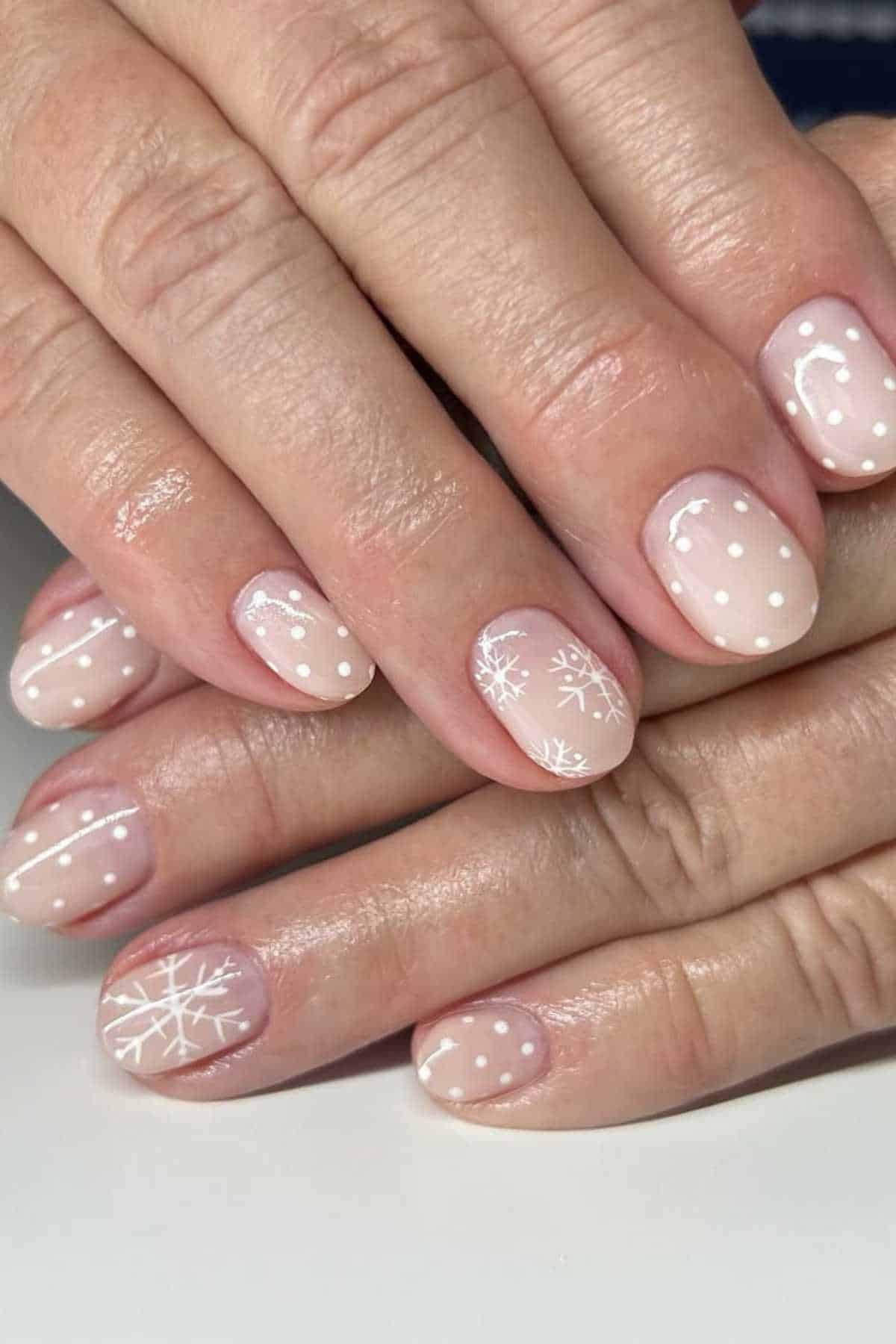 Hands with short, rounded nails painted in a nude shade, featuring white polka dots and snowflake designs on select nails.