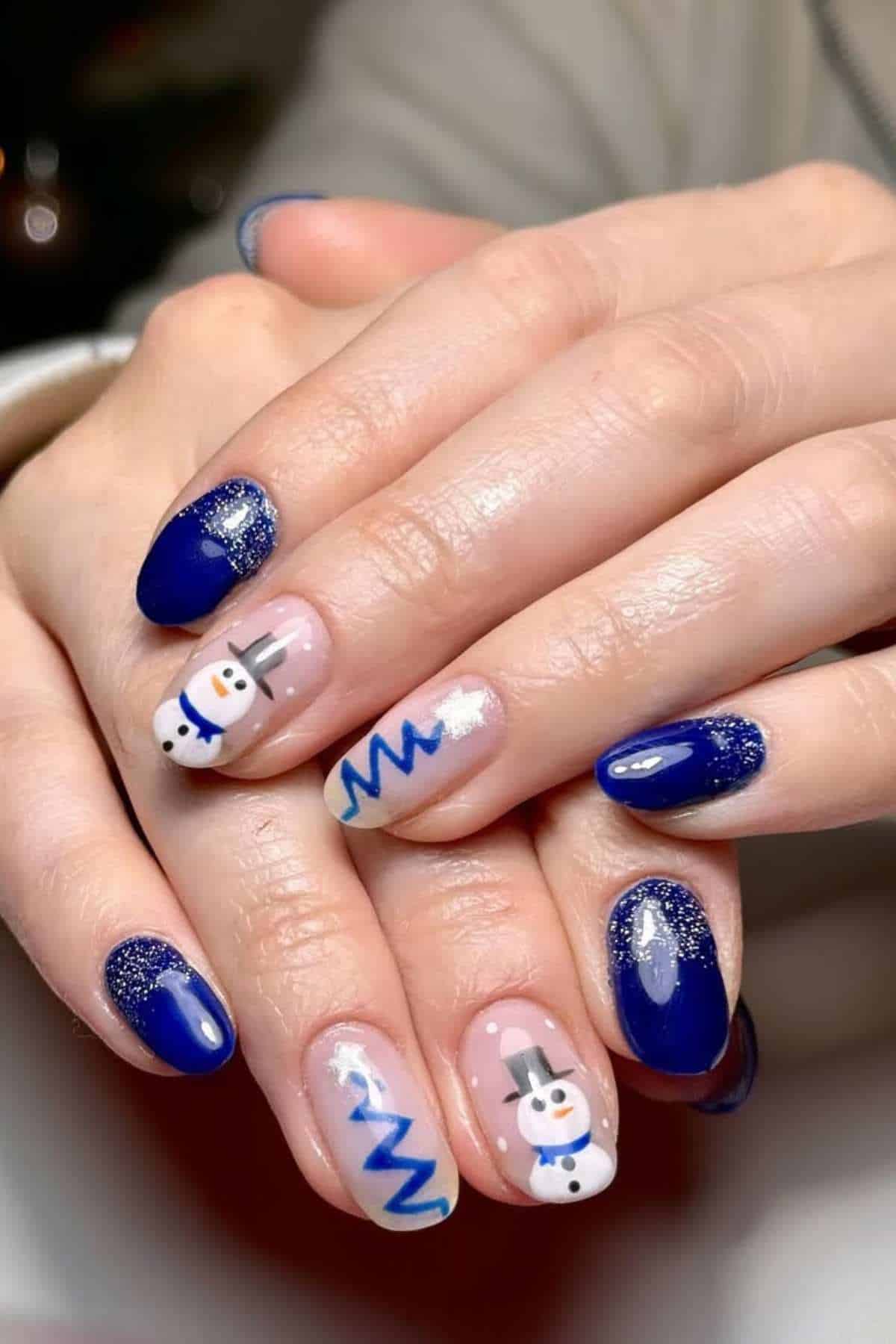 Hands with blue and glittery nail polish, featuring snowman and Christmas tree designs on accent nails.