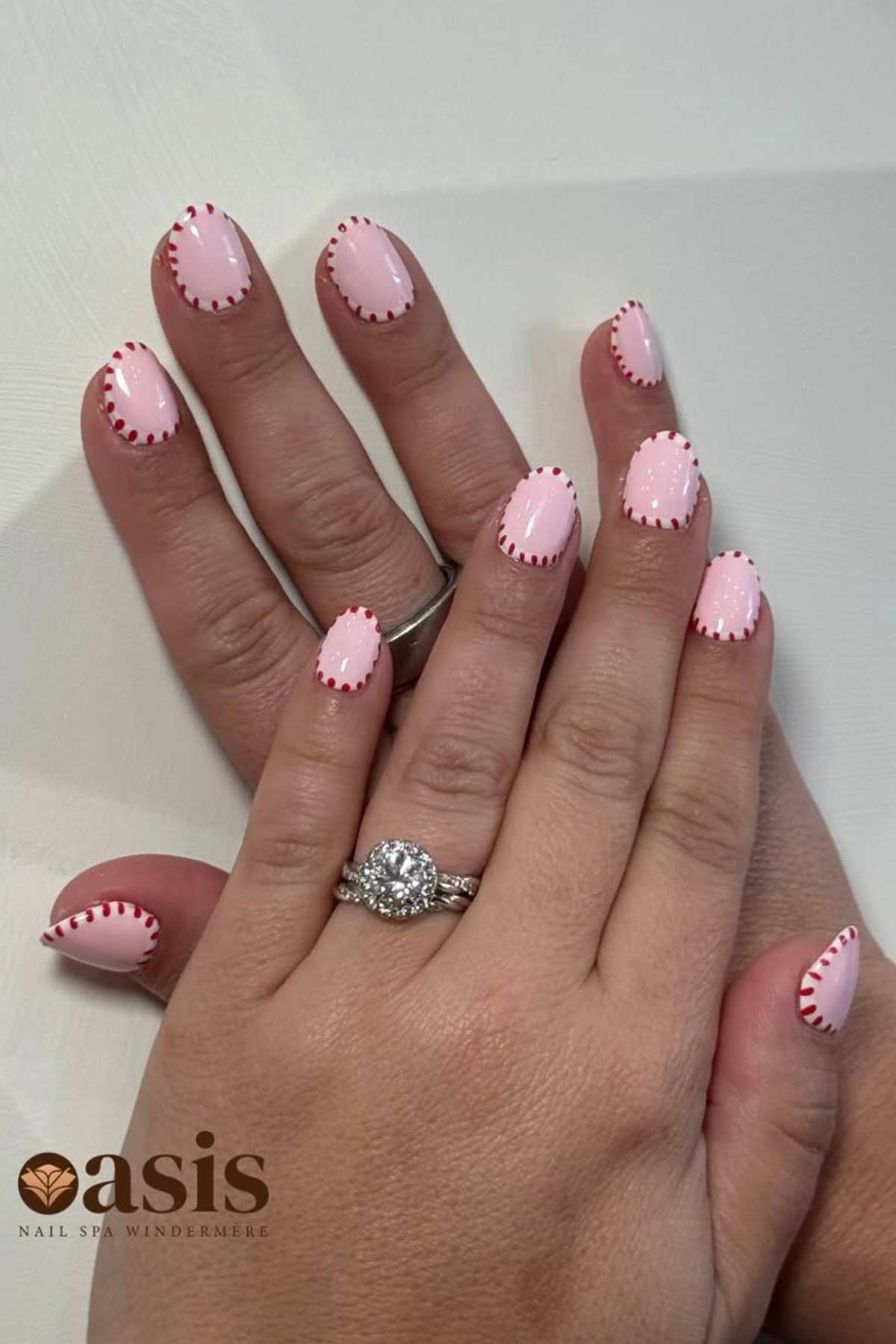 A hand with light pink nails featuring a stitched red border design, displaying two rings, including a diamond ring. The Oasis Nail Spa logo is visible in the corner.