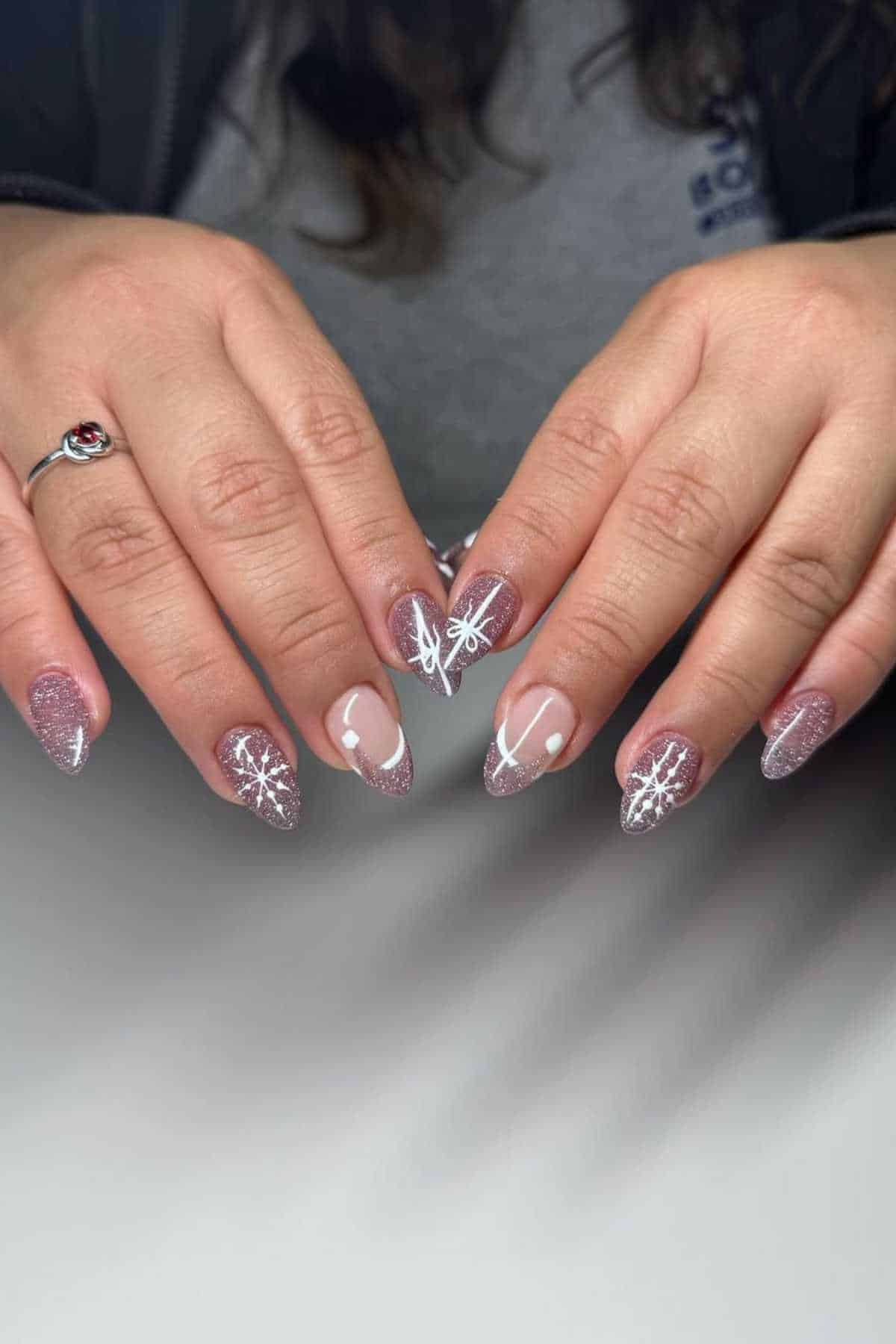 Hands with long, almond-shaped nails featuring pink glitter polish and white snowflake designs, placed on a white surface.
