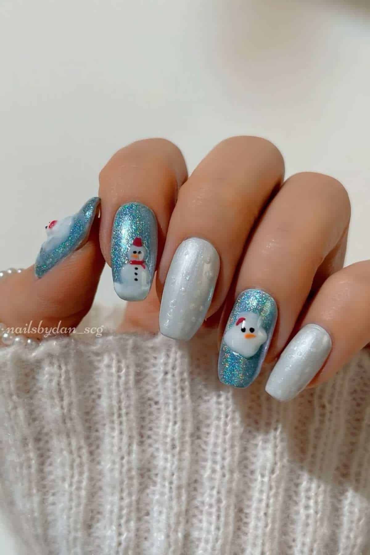 A hand with winter-themed nail art featuring silver, blue glitter, and snowmen designs, displayed against a white knit fabric background.