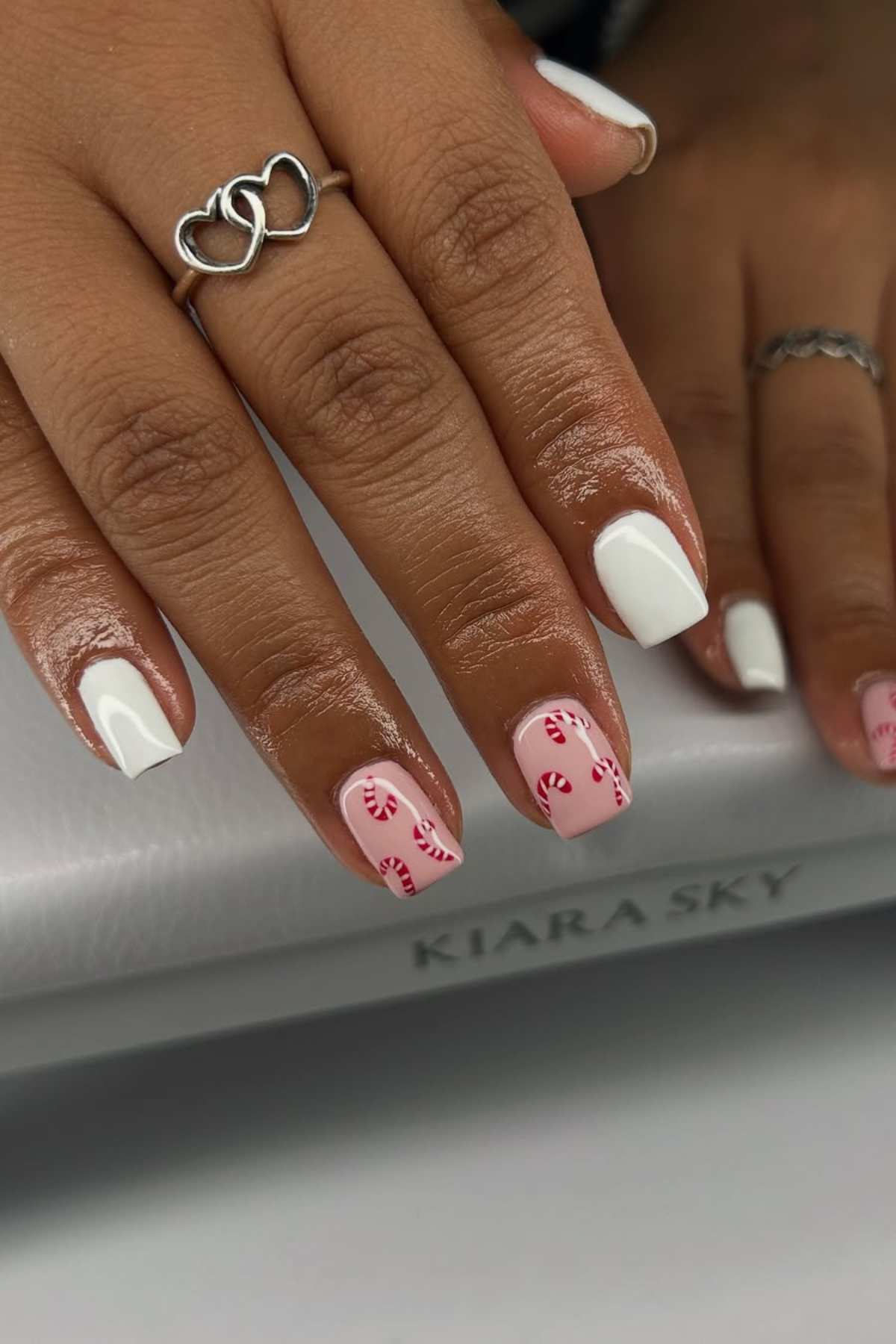 A hand with white and light pink nail polish; two nails feature red candy cane designs. The person wears two silver rings.
