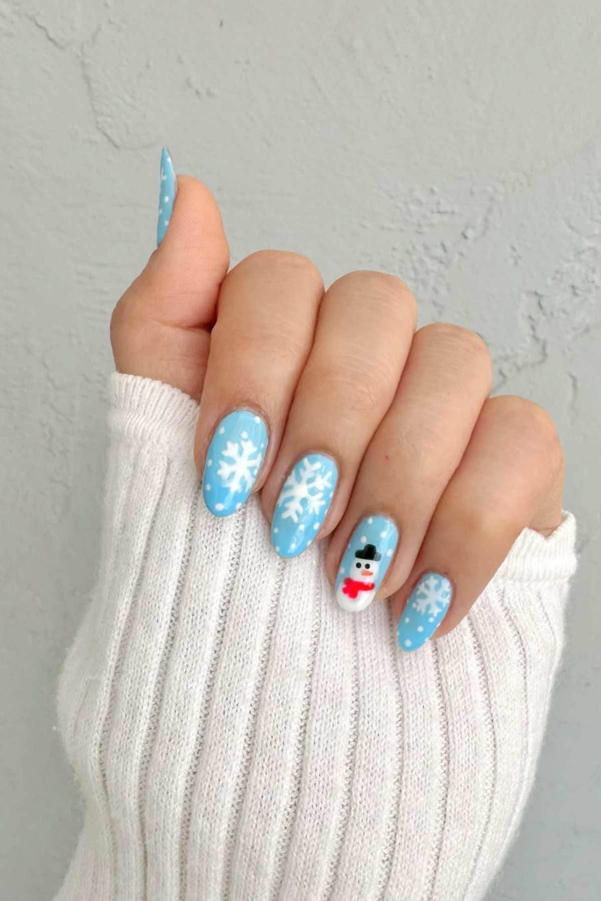 A hand with blue winter-themed nail art featuring white snowflakes, dots, and a small snowman design, shown against a light background.