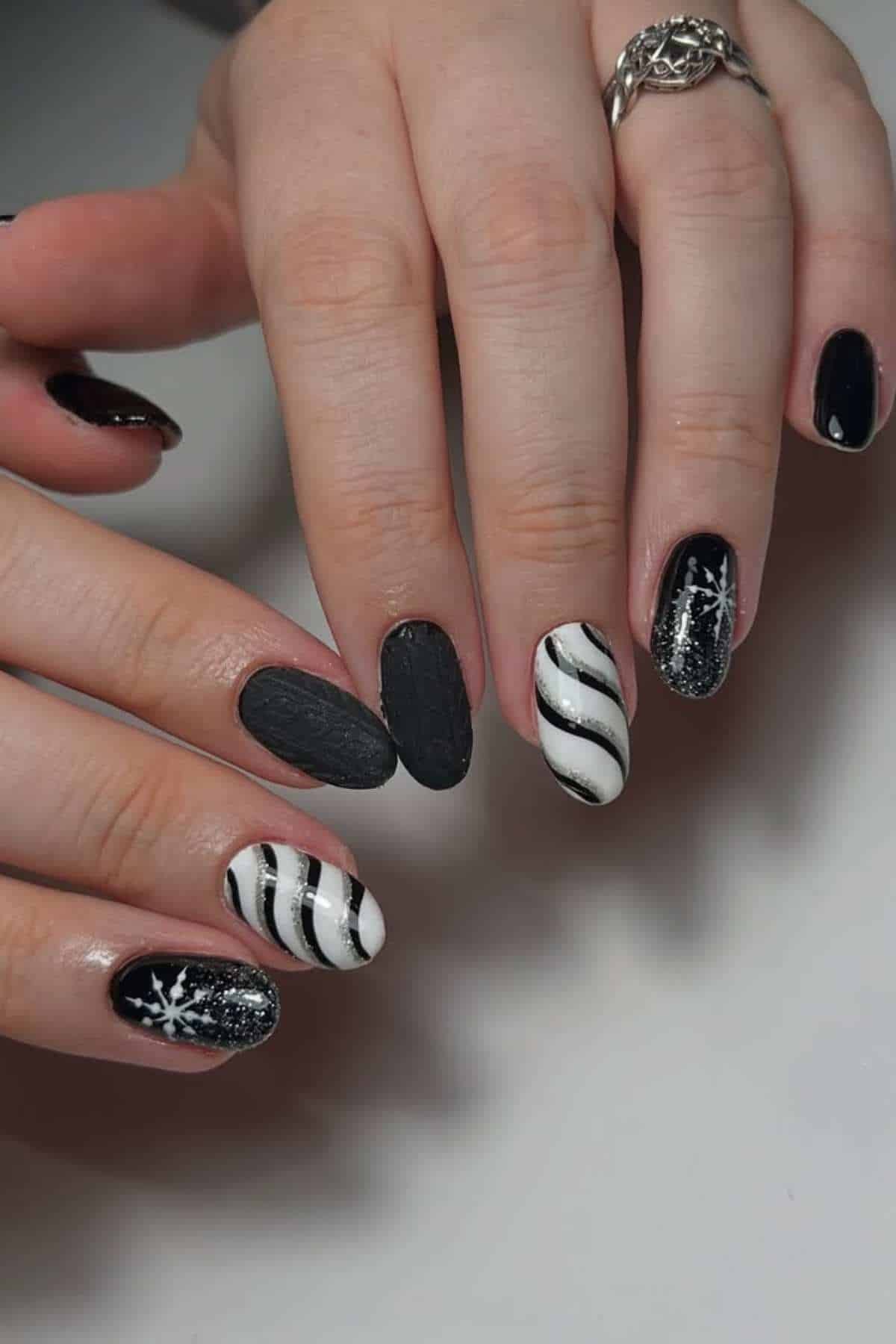 A hand with black, white, and glitter manicure featuring stripes, snowflakes, and matte finishes on almond-shaped nails.