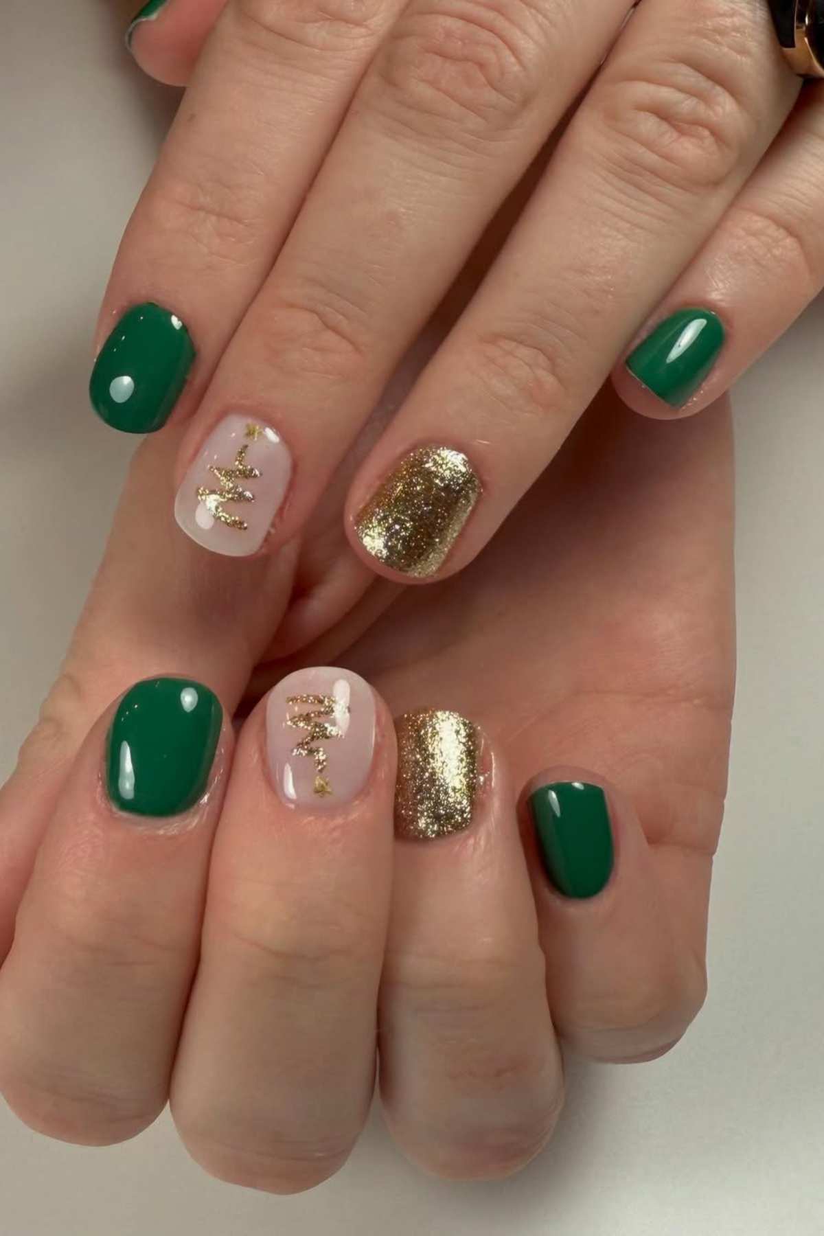A pair of hands with manicured nails featuring green polish, gold glitter accent nails, and clear nails with gold foil designs.