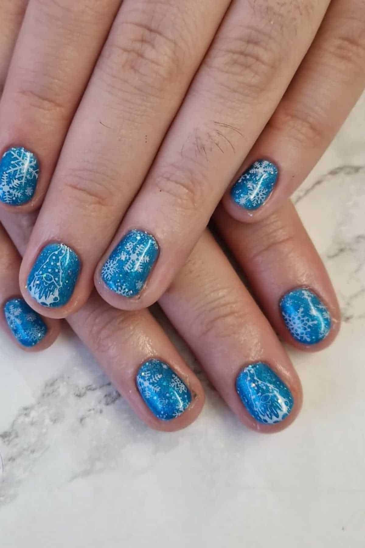 Hands with short nails painted blue, featuring white snowflake and gnome designs, placed on a marbled surface.