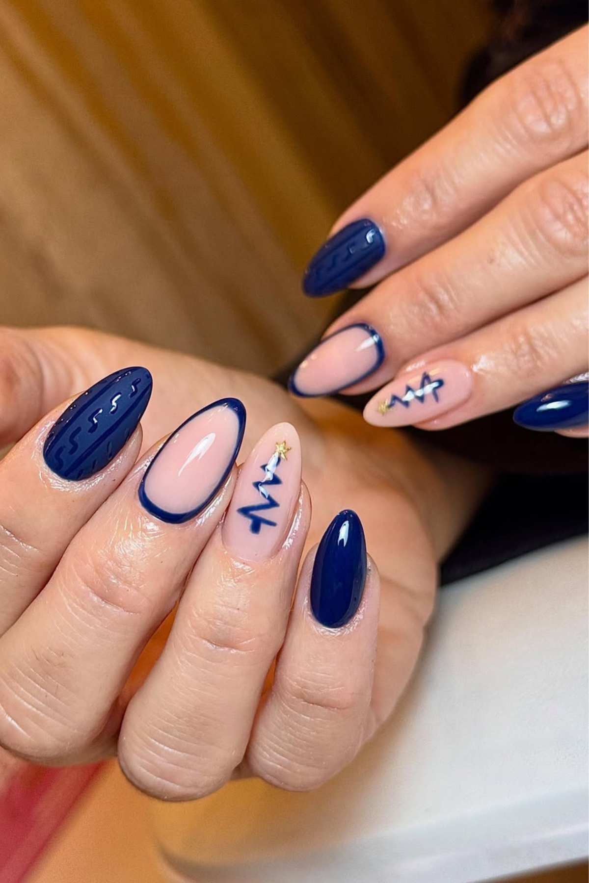Hands with almond-shaped nails featuring a blue and nude color scheme, some with glossy blue polish, others with nude base and blue Christmas tree designs.