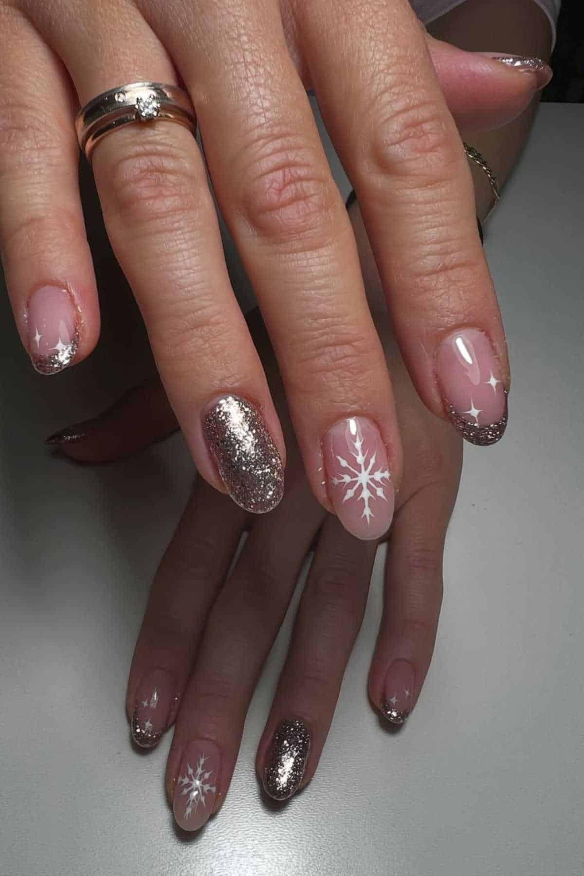 A hand with manicured nails featuring pink polish, silver glitter accents, and white snowflake designs on two nails, resting on a white surface.