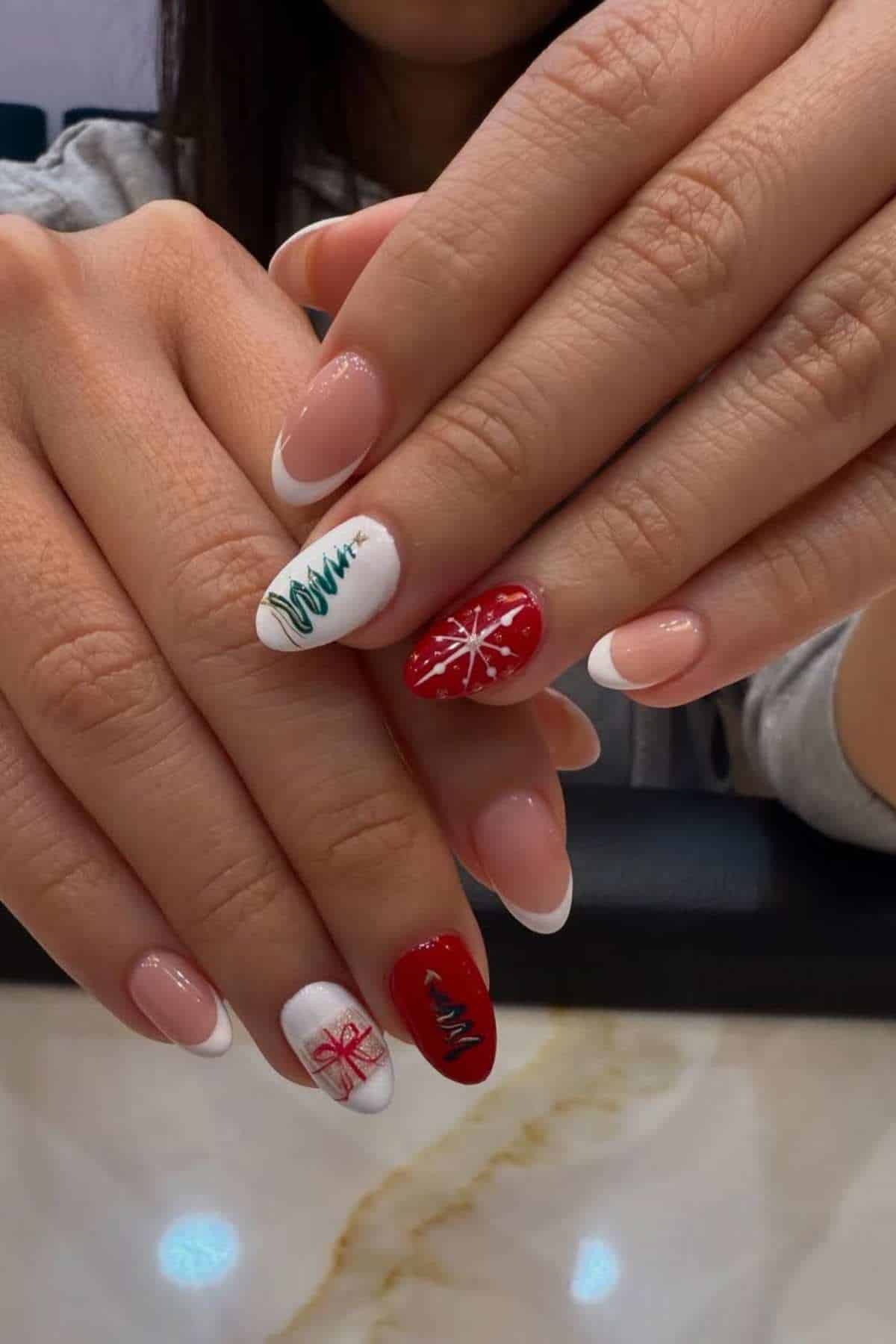 A person with French manicure nails; some nails are painted red or white with Christmas-themed designs including a tree, snowflake, and gift box.