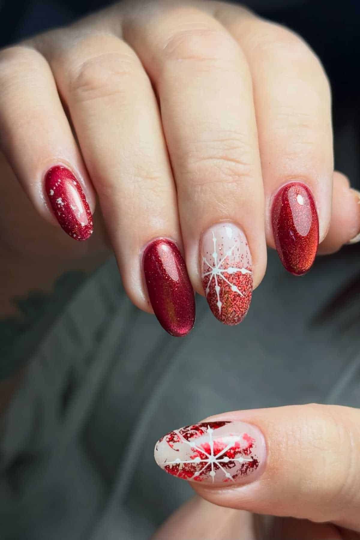 A hand with red glittery nails, some featuring white snowflake designs and gradient red tips, holding another hand with a similar festive manicure.