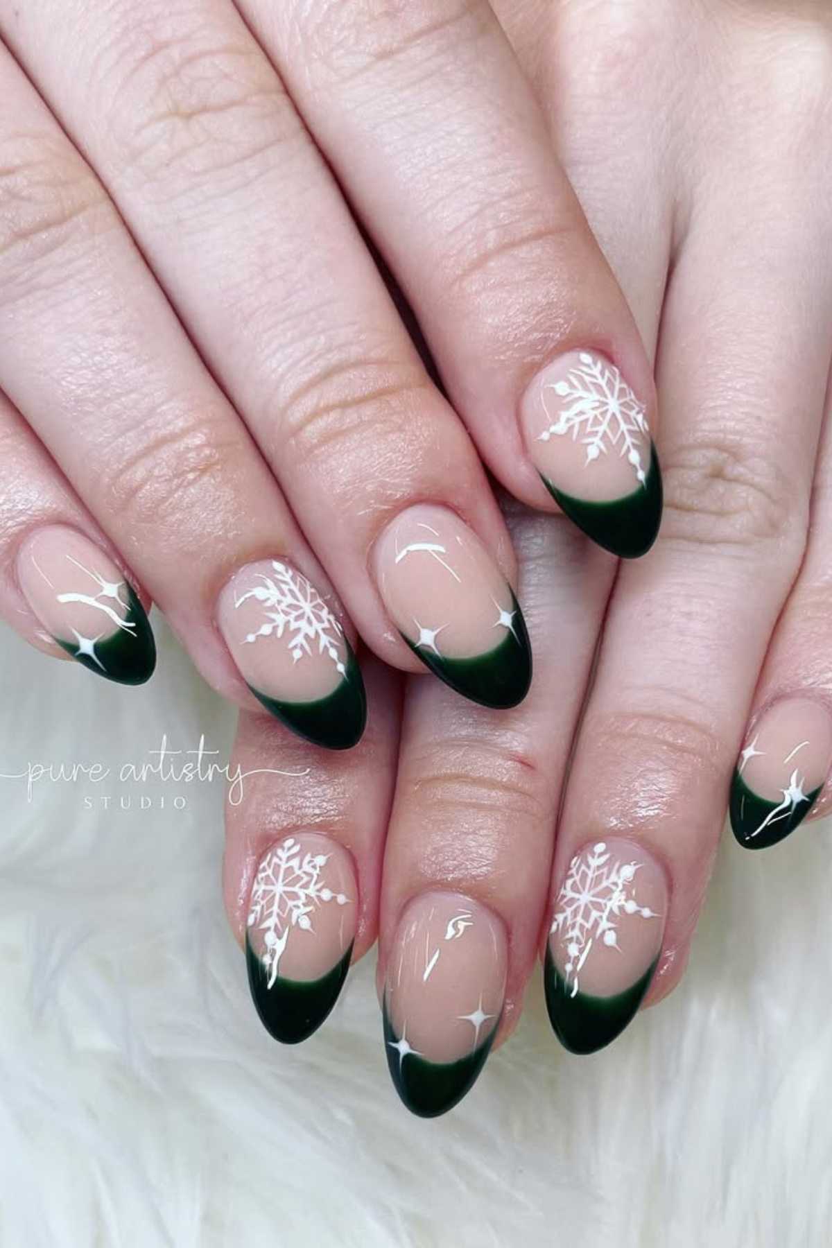 Hands with almond-shaped nails featuring dark green French tips and white snowflake designs on a light, neutral base.