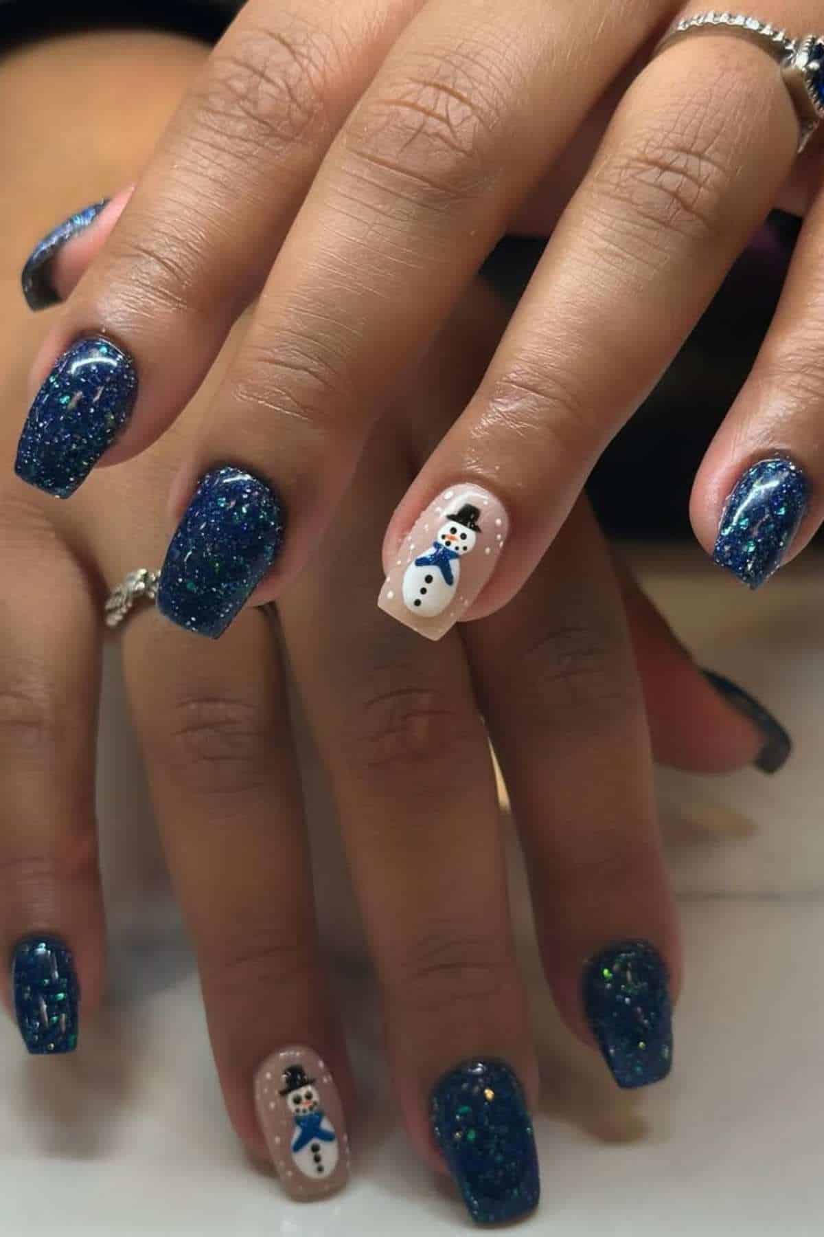 Hands with blue glittery nail polish and accent nails featuring a snowman design on a white background.
