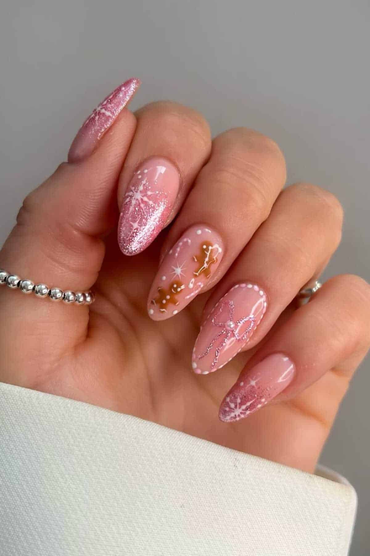 A hand with long, almond-shaped nails painted pink, featuring glitter, bows, snowflakes, and gingerbread man nail art.