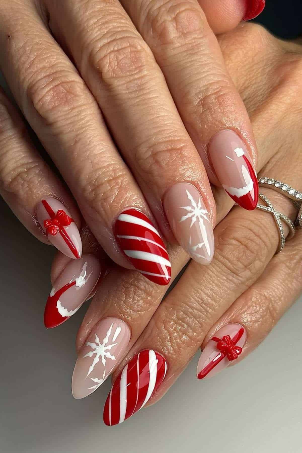 A hand with almond-shaped nails featuring red, white, and nude Christmas-themed designs, including stripes, snowflakes, bows, and gift wrap patterns.