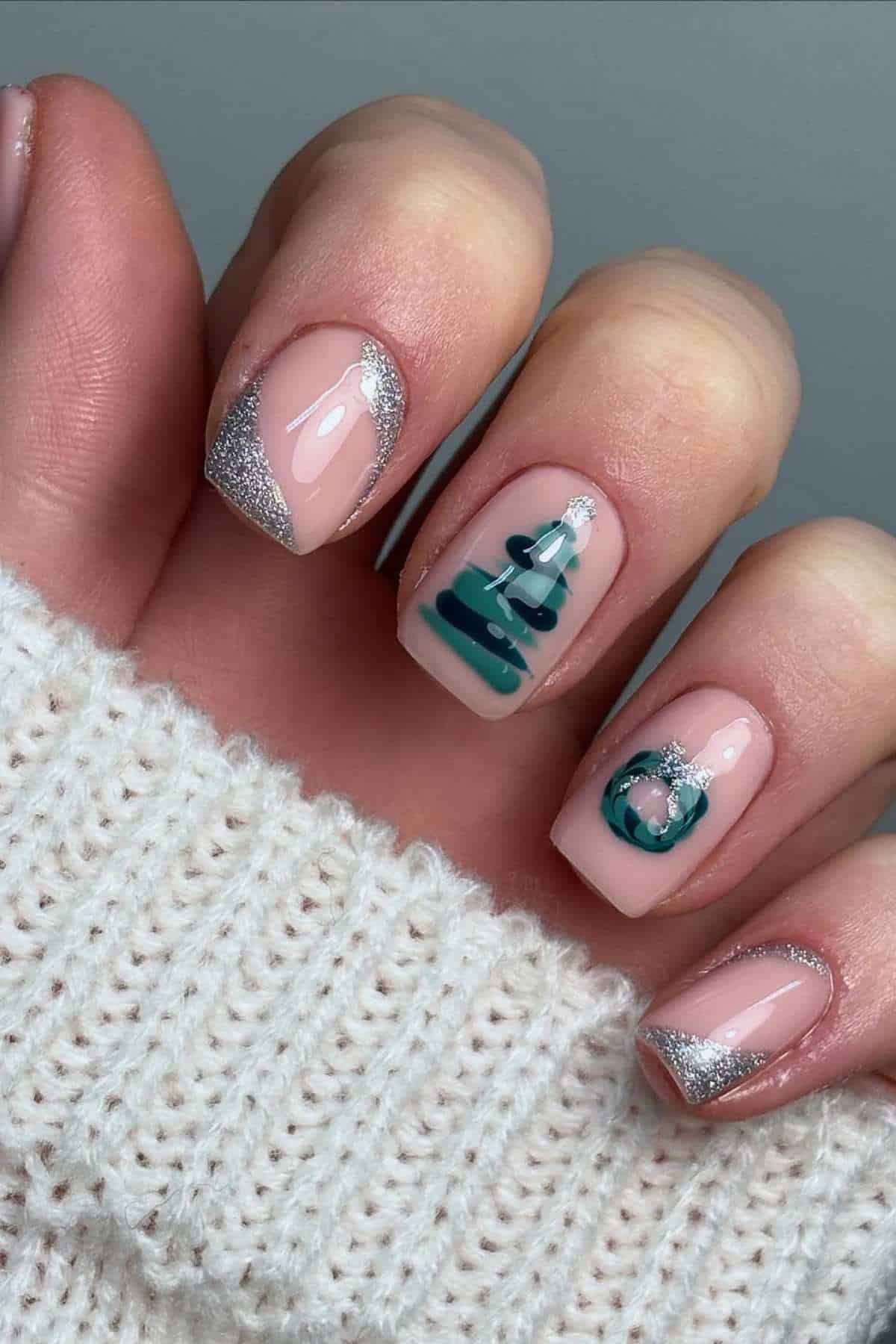 A hand with nude-colored nails featuring silver glitter tips and accent nails with a green Christmas tree and a green ornament design. The hand is partially covered by a white knit sweater.