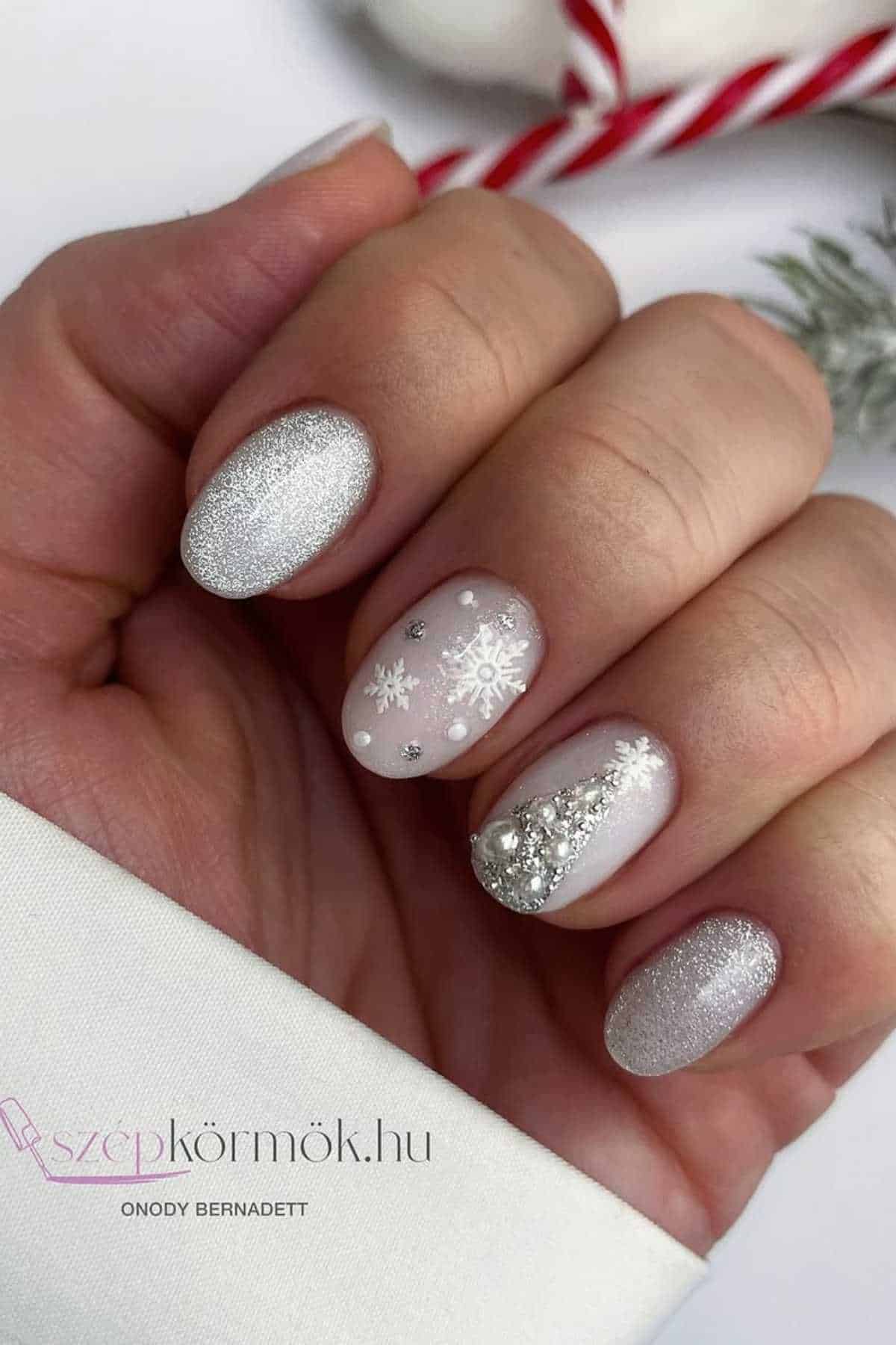 A hand with silver and light pink winter-themed nail art, featuring snowflakes and rhinestone decorations on the ring and middle fingers.