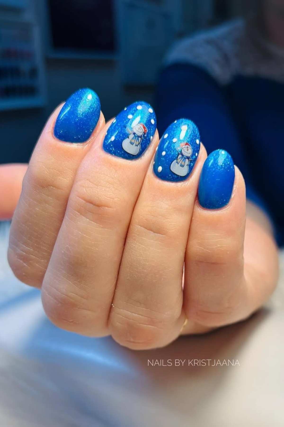 A hand with blue painted nails featuring snowman designs and small white dots, photographed indoors.