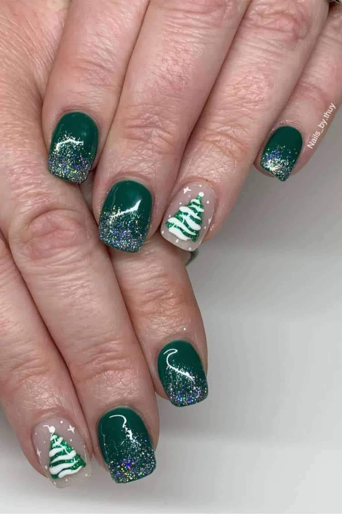 Hands with green glittery nail polish, featuring white Christmas tree designs on the ring finger nails, against a plain background.