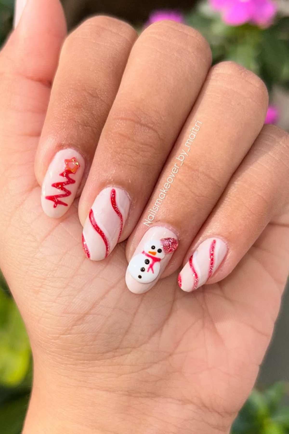 A hand with festive nail art featuring red glittery lines, a Christmas tree on one nail, and a snowman design on another, with a pink floral background.