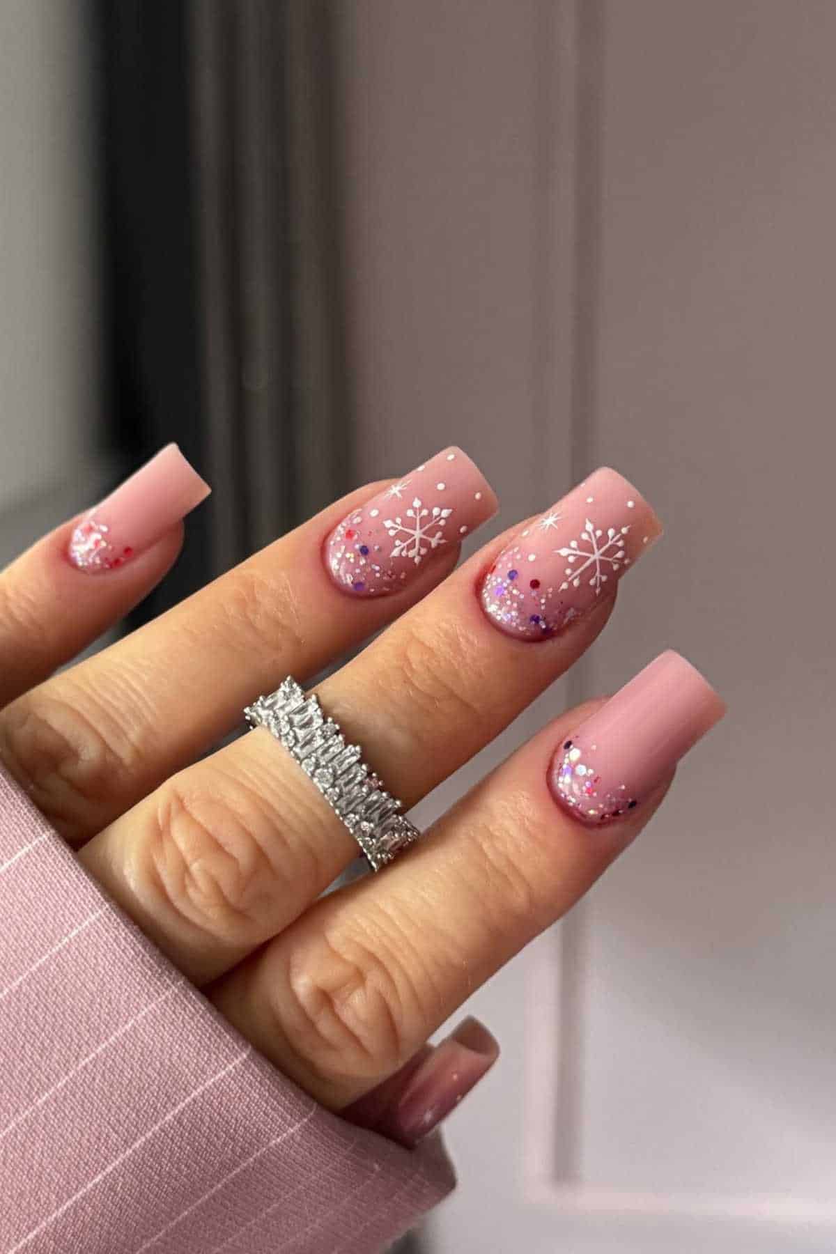 A hand with long, square-shaped pink nails featuring white snowflake designs and glitter accents, with a silver ring on the ring finger.