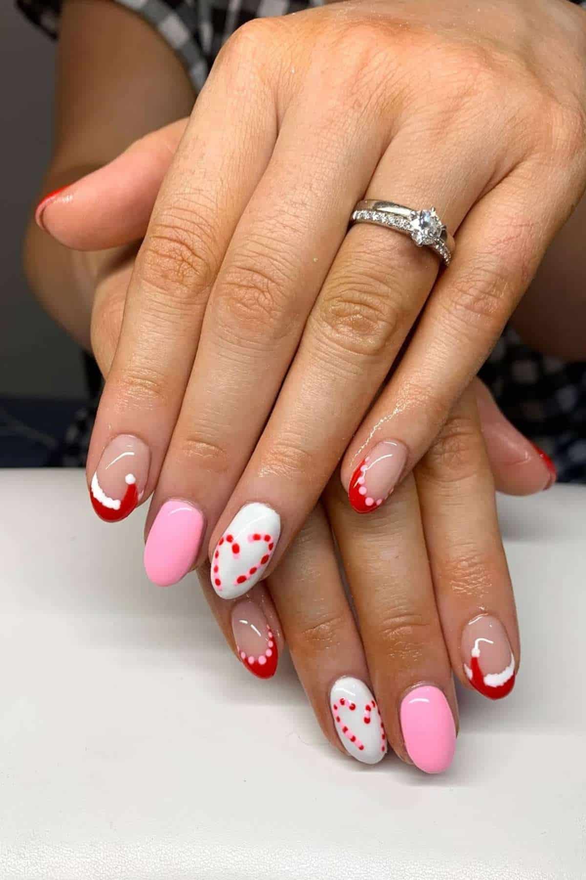 Hands with manicured nails featuring pink polish, red French tips, and red dotted designs. Person is wearing a diamond ring on their left hand.