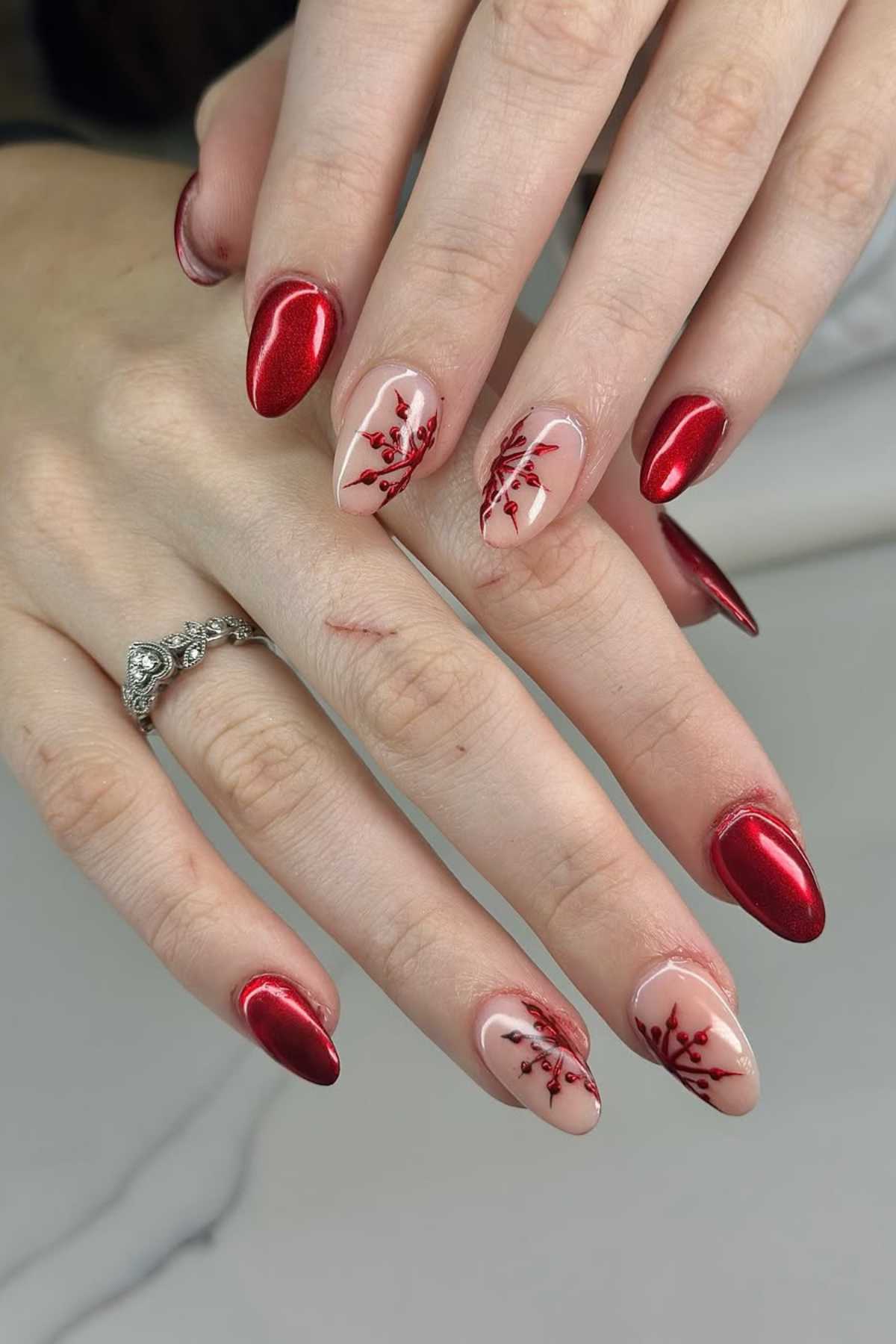 A hand with almond-shaped red metallic nails and floral branch designs on two accent nails. A silver ring is worn on one finger.