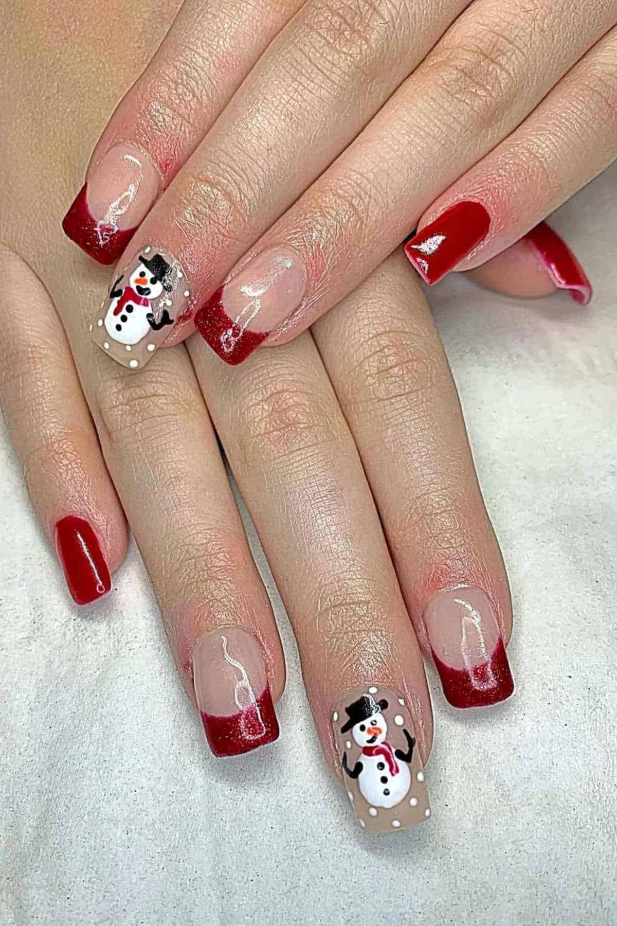 Hands with red French tip manicure; two nails feature snowman designs with white dots, resembling snow.