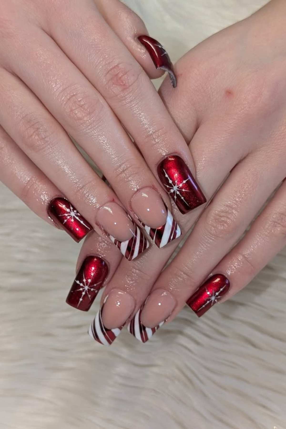 Hands with acrylic nails featuring a mix of red metallic polish with white snowflake designs and nude polish with white and red candy cane stripe tips, set on a light fur background.