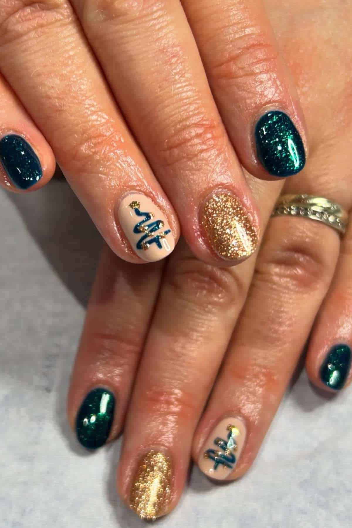 Close-up of hands with short, oval nails painted in green glitter, gold glitter, and beige polish, with blue and gold tree designs on two accent nails.