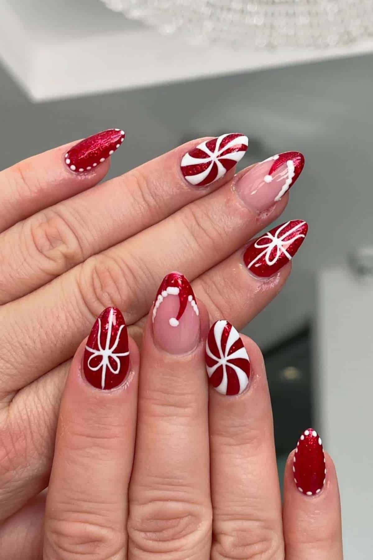 A hand with almond-shaped nails painted red and decorated with white swirls, dots, and lines in holiday-themed designs.
