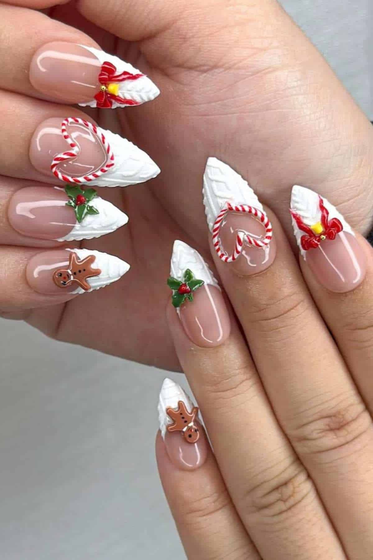 Hands with long, almond-shaped Christmas-themed nail art featuring white textured tips, gingerbread men, holly leaves, candy canes, and red ribbons on a nude base.