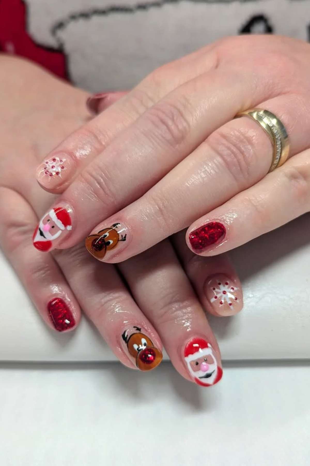 A hand with festive nail art featuring Santa Claus, Rudolph the Red-Nosed Reindeer, red glitter, and snowflakes on a light background.