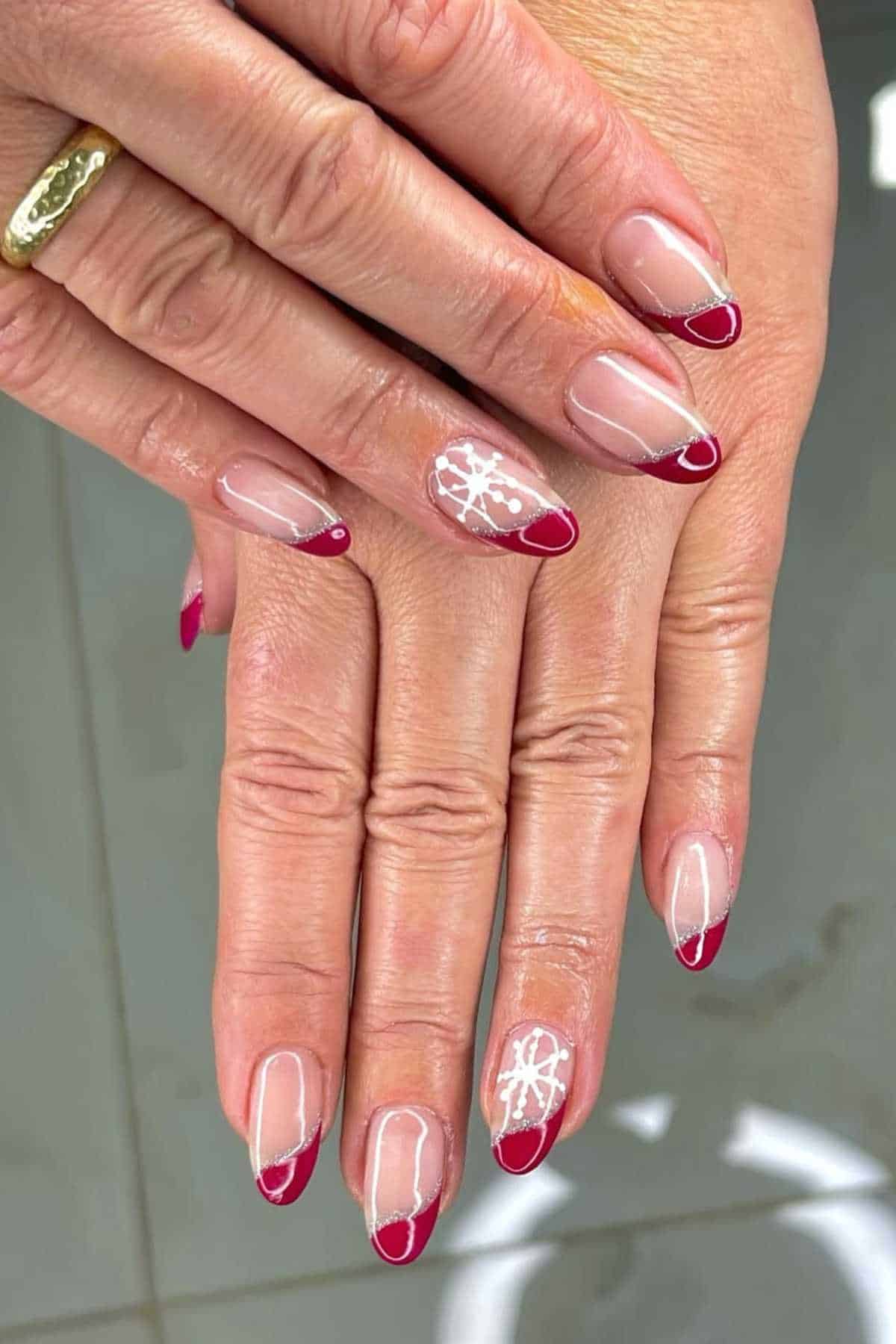 Hands with almond-shaped nails featuring red French tips, one accent nail with white floral design, and a gold ring on one finger.