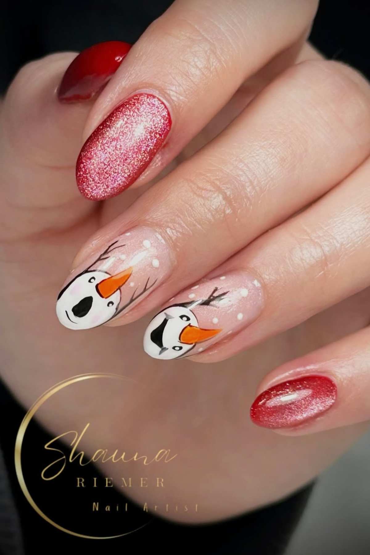A hand with red glittery nails and two accent nails featuring snowman faces with orange noses and antlers on a nude background.