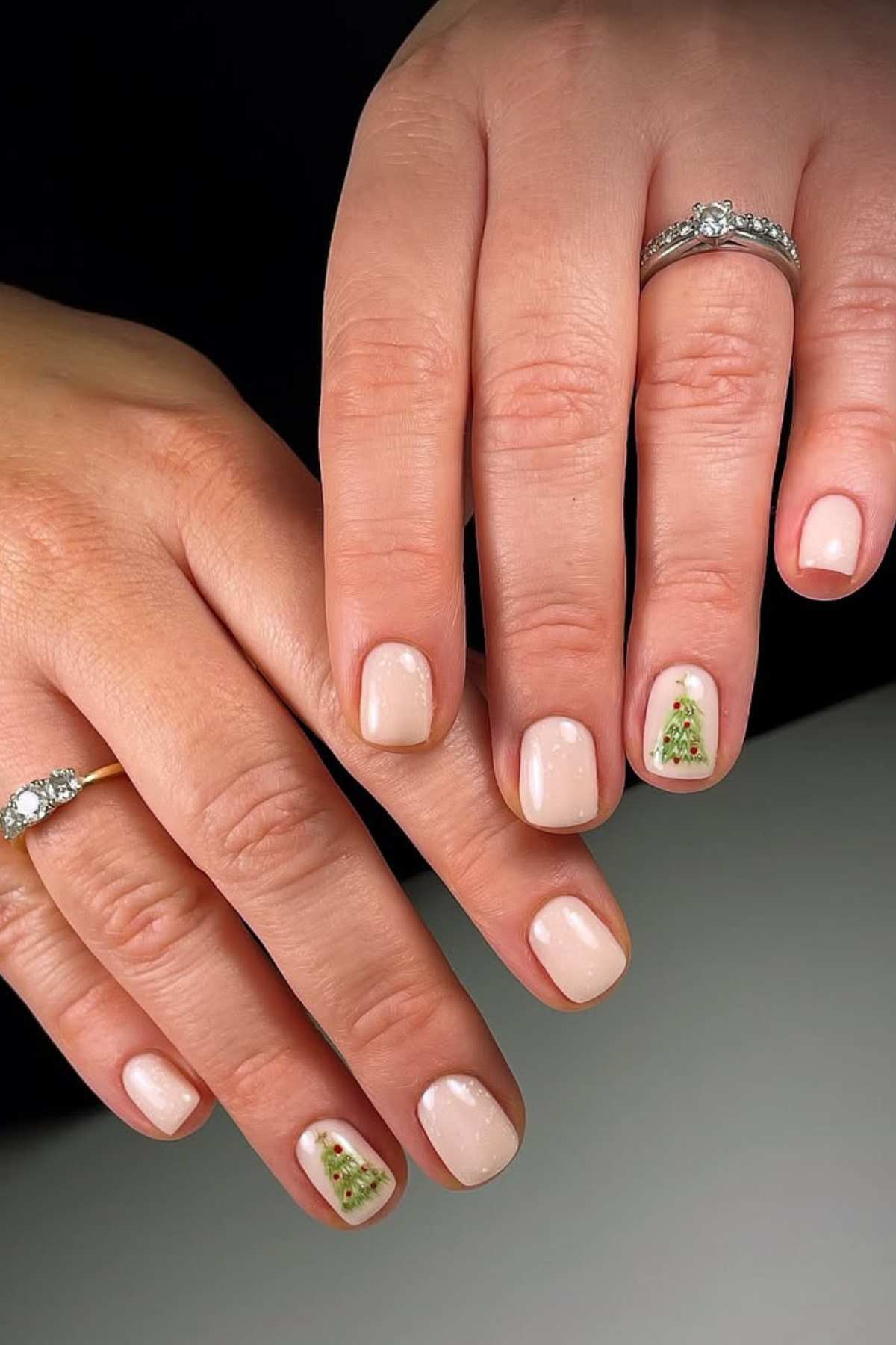 Two hands with short, pale pink nails; each ring finger features a small green Christmas tree design with red accents.