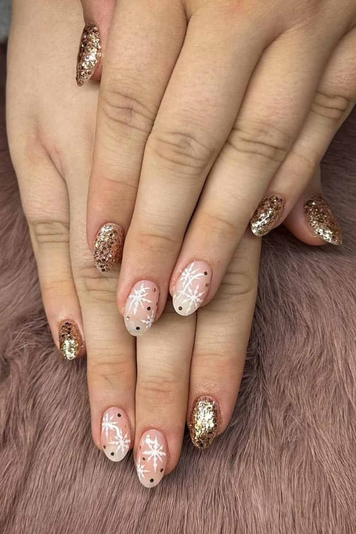 Hands with almond-shaped nails featuring gold glitter polish on some nails and nude polish with white snowflake designs and black dots on others, resting on a soft textured surface.