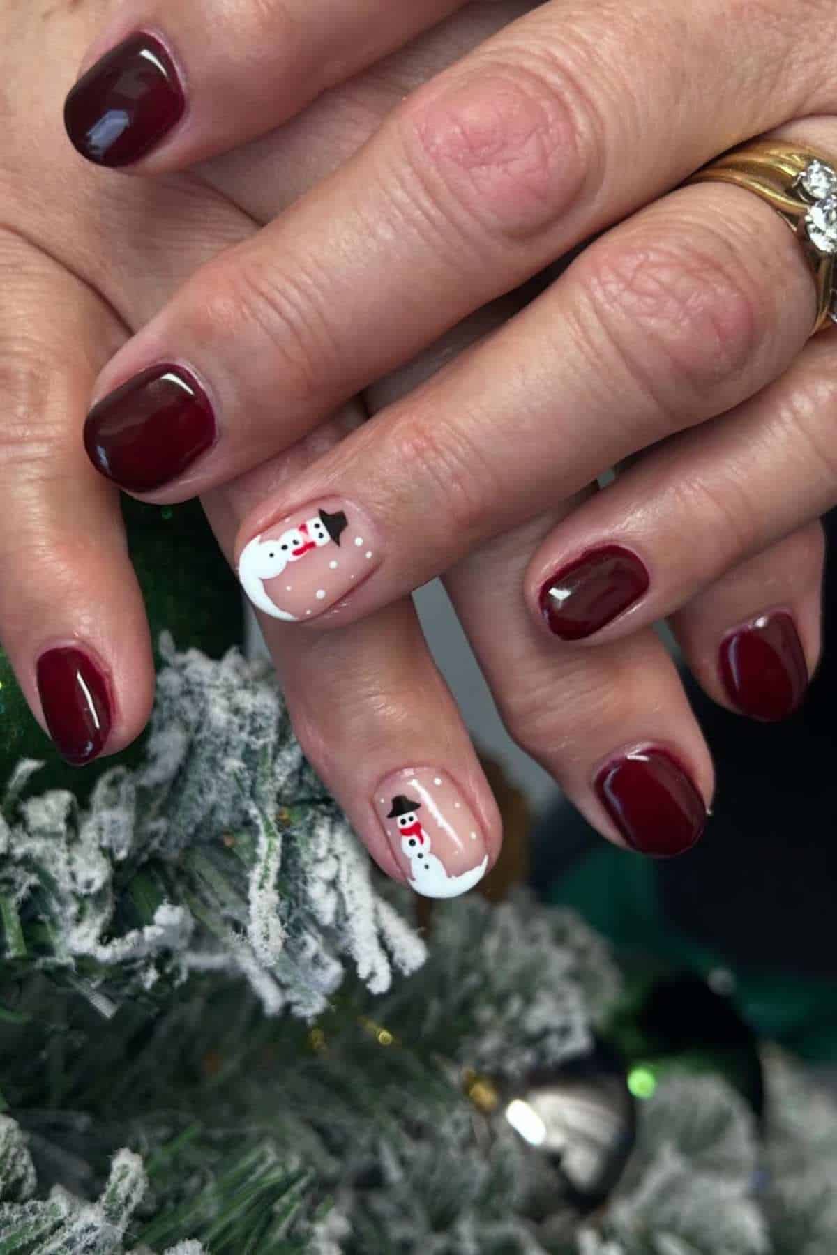 A hand with dark red manicured nails and an accent nail on each hand featuring a painted snowman design.