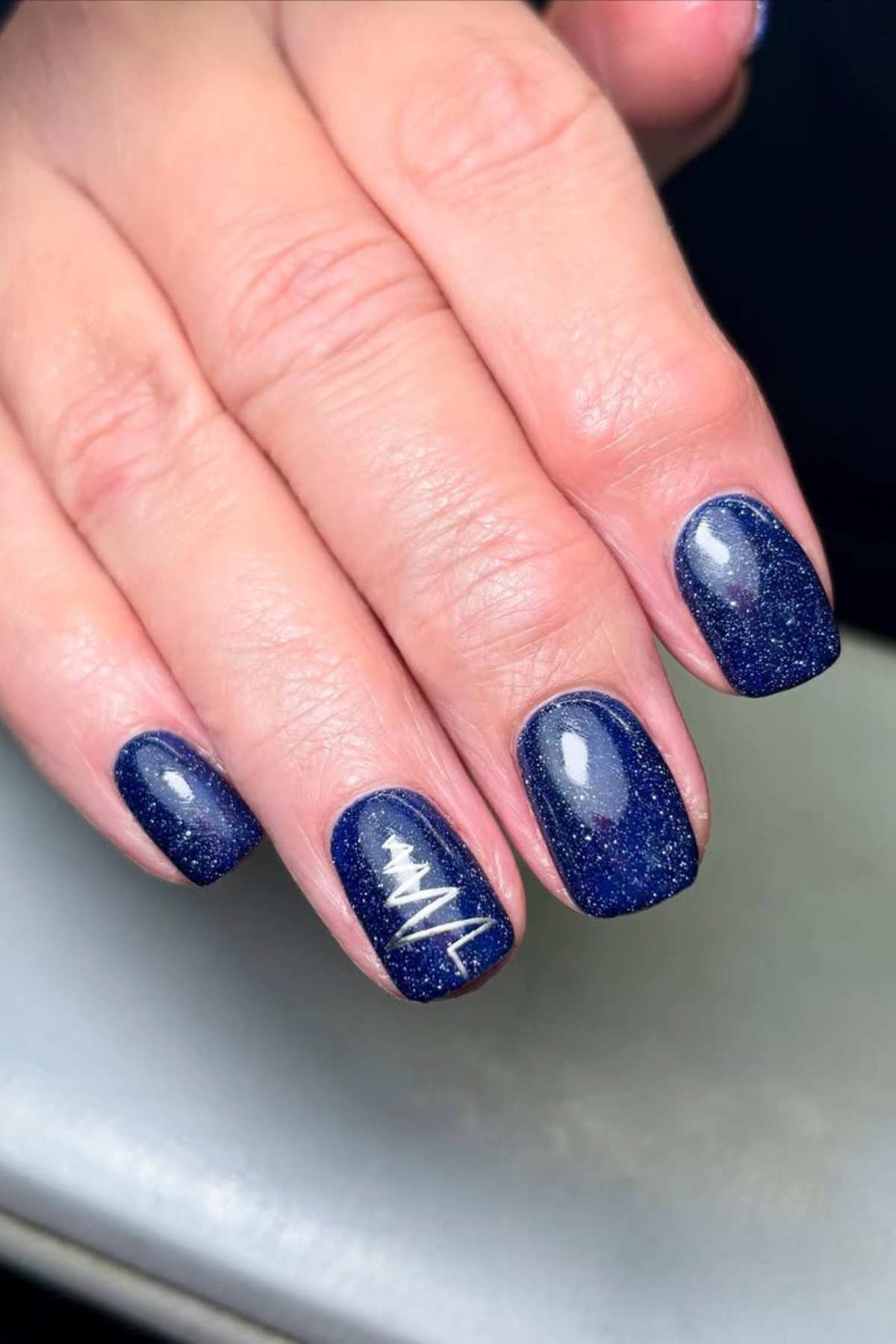 A hand with short, square-shaped nails painted dark blue with a glittery finish; the ring fingernail features a white heartbeat line design.