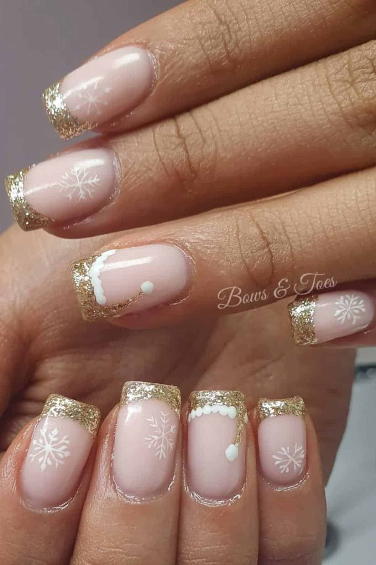 Close-up of hands with nude nails featuring gold glitter French tips, white snowflake designs, and small white decorative accents.