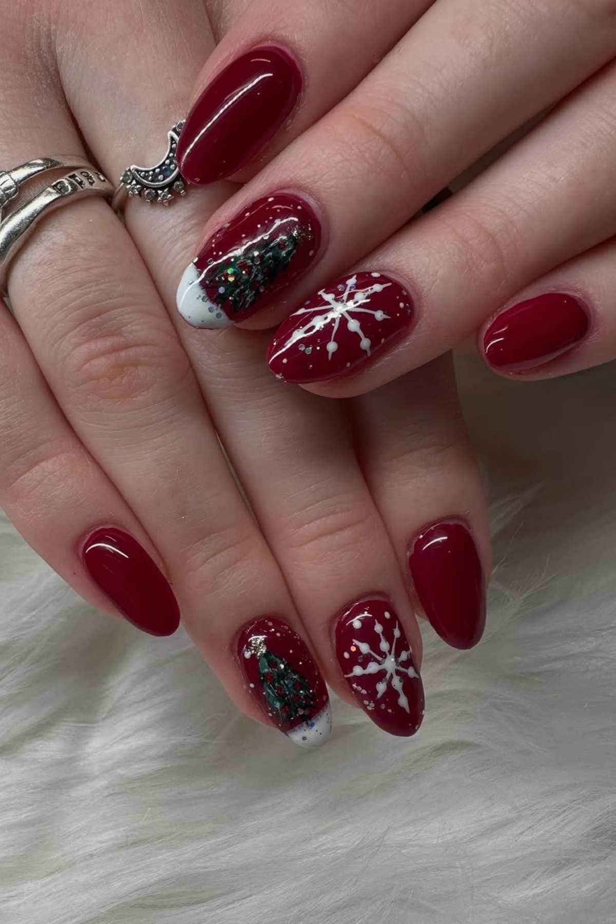 Hands with red Christmas-themed manicure featuring white snowflakes and a green Christmas tree design on two nails.