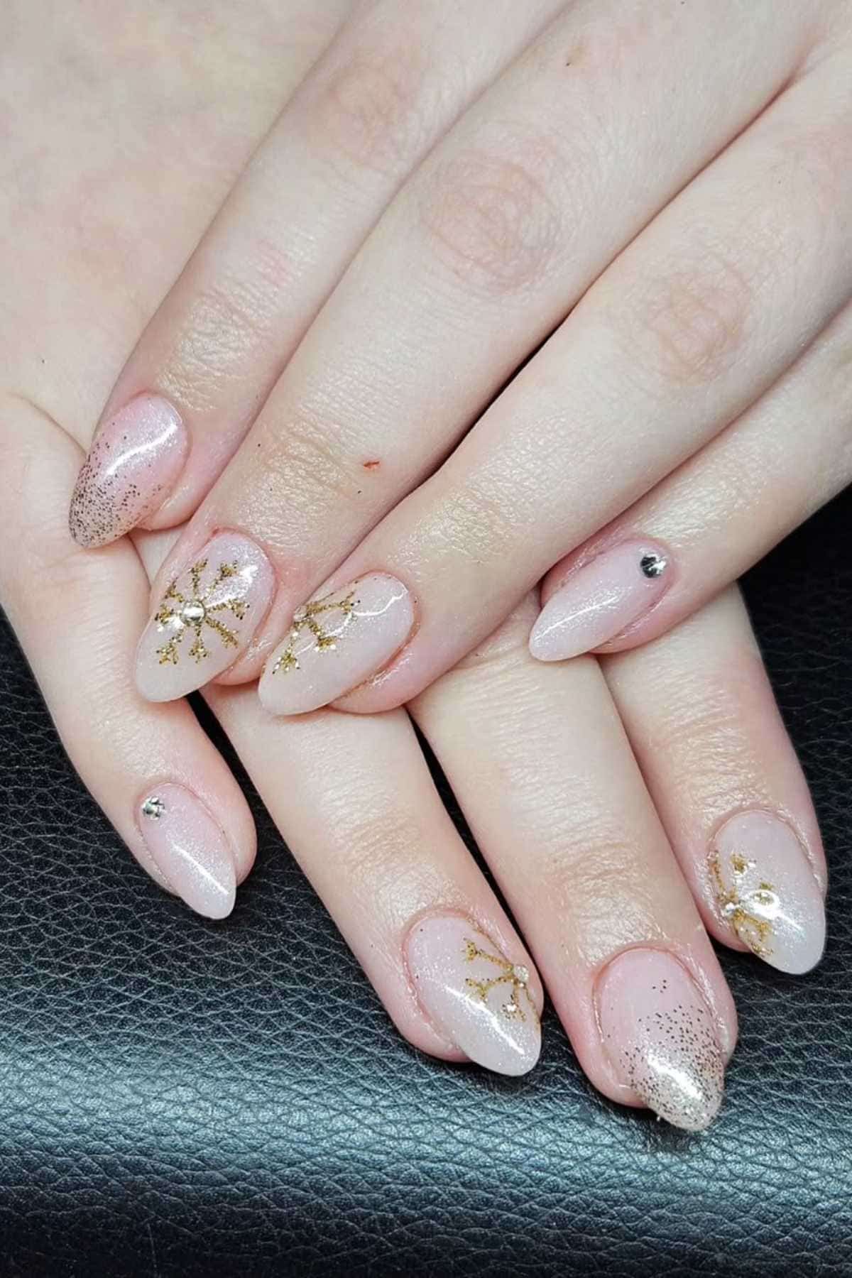 Close-up of hands with almond-shaped nails painted in a pale pink shade, decorated with gold floral designs, glitter, and small rhinestones.