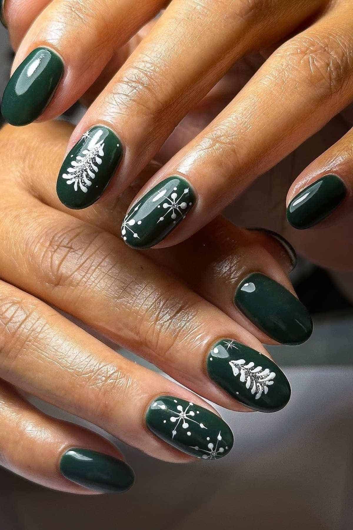 Hands with dark green nail polish featuring white holiday-themed designs, including snowflakes and pine trees, on some nails.