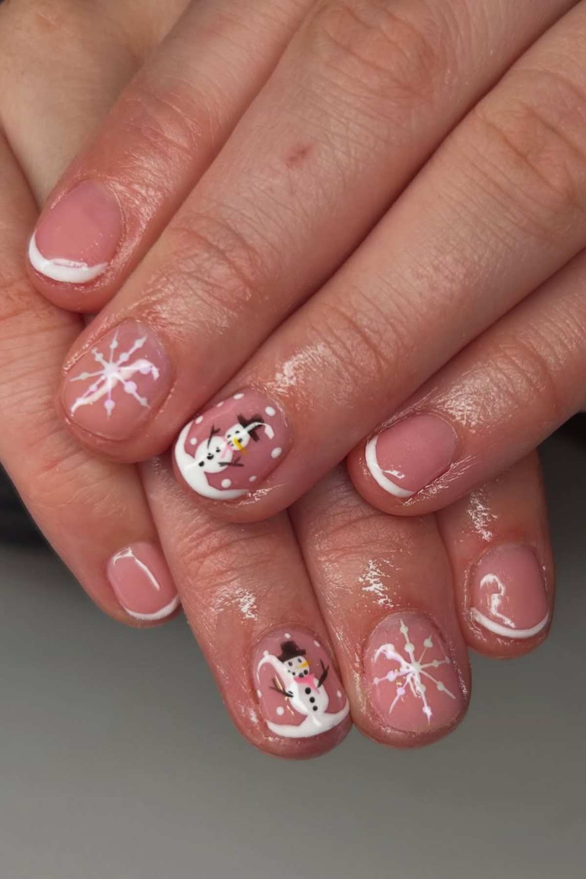 Close-up of hands with winter-themed nail art featuring white snowflakes and snowmen on a nude pink base, with a glossy finish.