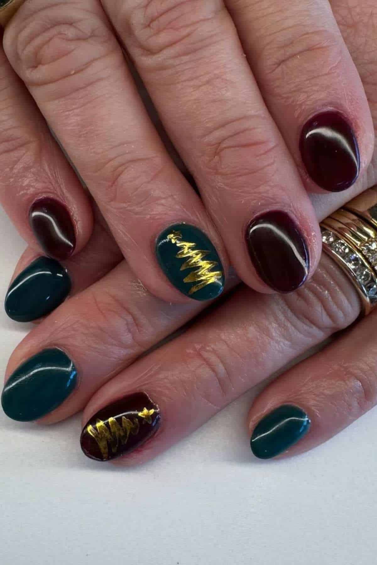 Hands with manicured nails painted in dark green and burgundy; two nails feature gold Christmas tree designs. Rings are worn on two fingers.
