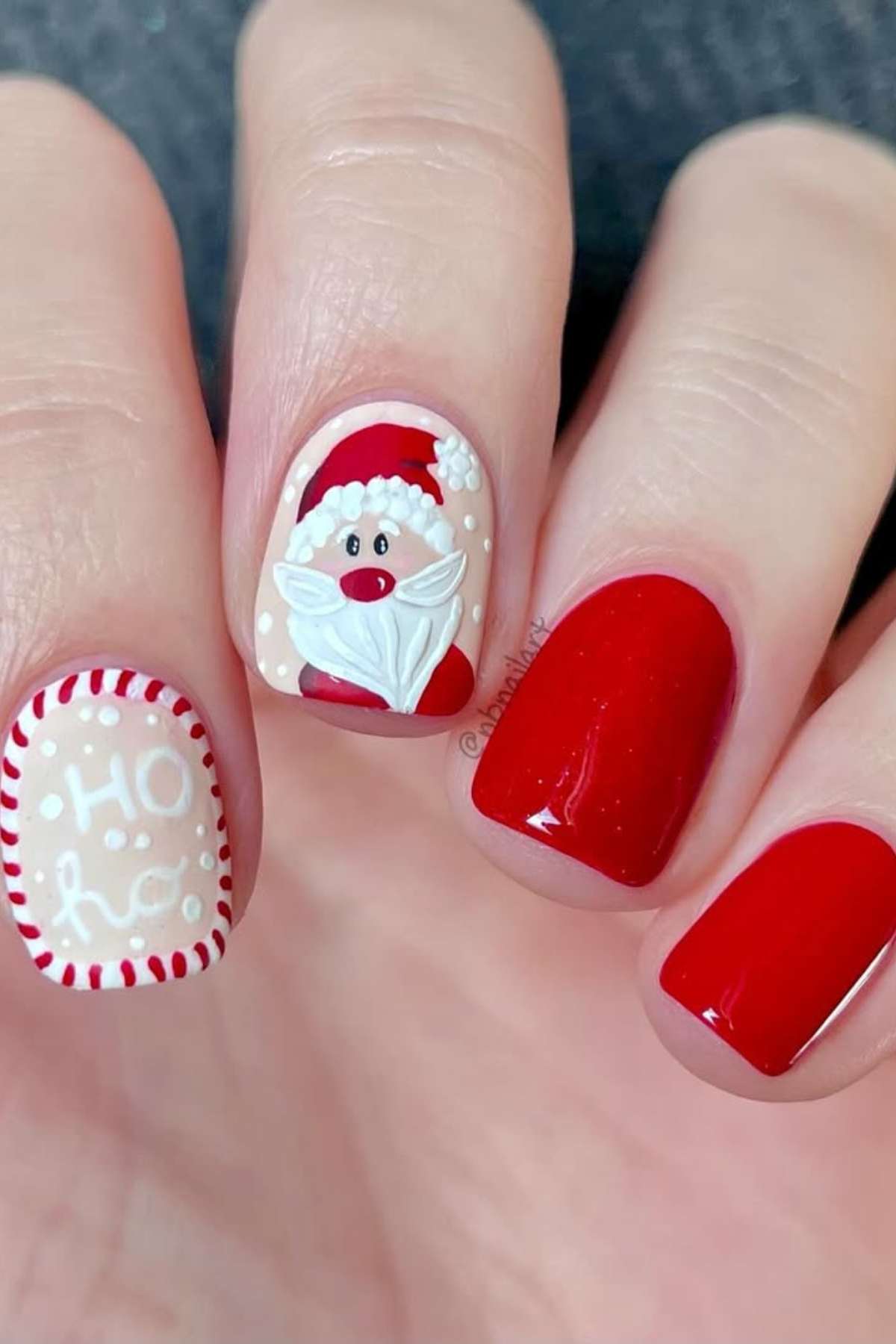 Close-up of a hand with Christmas-themed nail art featuring red polish, a Santa Claus face, and "Ho Ho" text with decorative white and red details.