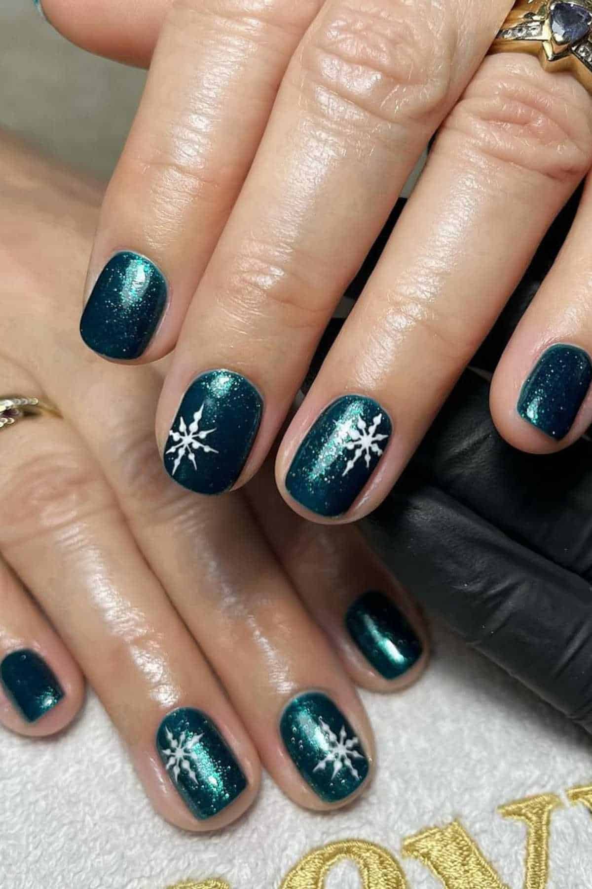 Hands with short, dark teal glittery nails, some with white snowflake designs. Person is wearing gold and silver rings.