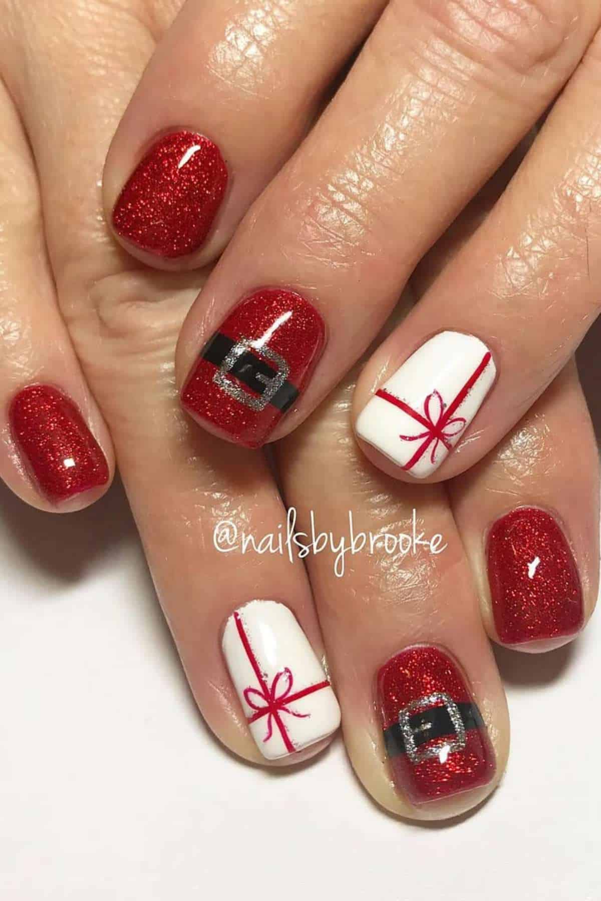 A hand with red glitter nail polish and accent nails featuring white polish with red ribbon designs and Santa belt art.