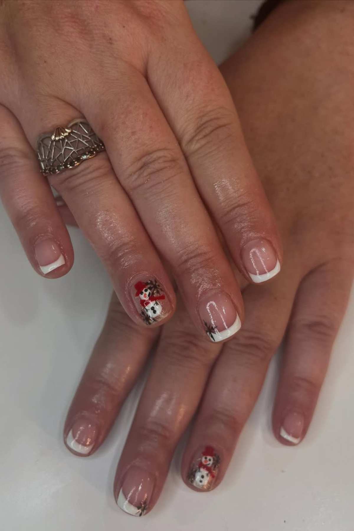 Two hands with French manicured nails, featuring snowman nail art on the ring fingers and a silver ring on one finger.